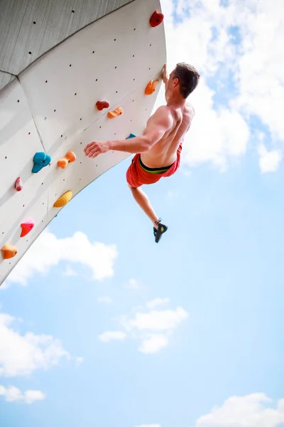 Foto von unten eines jungen athletischen Mannes, der an einer Wand übt, um gegen blauen Himmel mit Wolken zu klettern — Stockfoto