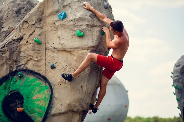 Foto do jovem desportista em calções vermelhos exercitando-se em pedra para subir contra o céu nublado — Fotografia de Stock