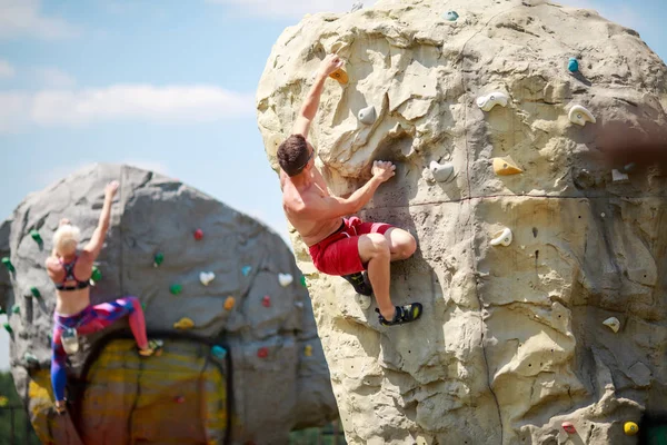 Photo from back of sports guy in red shorts and woman on workout at climbing boulders against blue sky with clouds — Stock Photo, Image