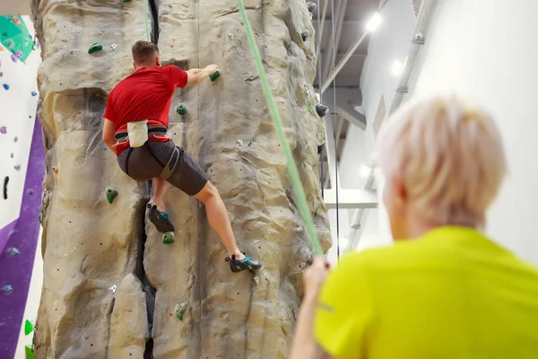 Foto von hinten: Sportler klettert über Kletterwand und Trainerin hält Sicherungsseil in der Hand — Stockfoto
