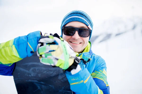 Retrato de hombre feliz en gafas de sol con snowboard en manos sobre fondo de montañas nevadas —  Fotos de Stock