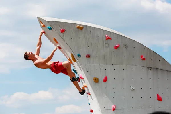 Foto eines jungen Sportlers in roten Shorts an der Wand zum Klettern vor blauem Himmel mit Wolken — Stockfoto