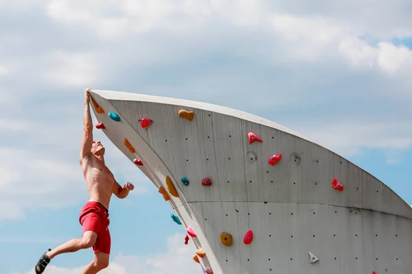 Photo of trained sports guy in red shorts hanging on wall for rock climbing against blue sky with clouds