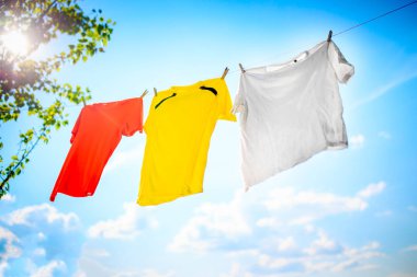 Photo of yellow, white and orange T-shirt hanging on rope against blue sky background with treetops. clipart