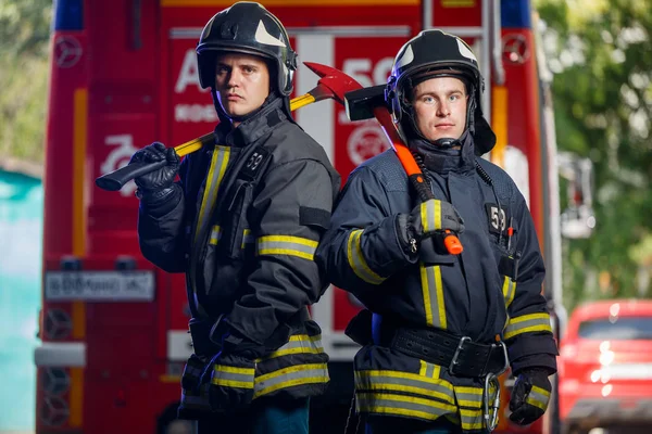 Foto de dos bomberos con hachas en las manos cerca del camión de bomberos — Foto de Stock