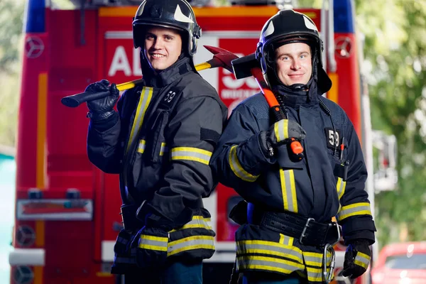 Foto av två unga brandmän med yxor i händerna nära brandbil — Stockfoto