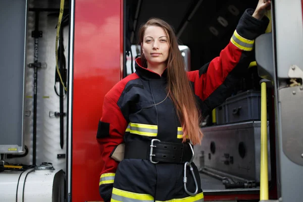 Foto de una joven bombera con el pelo largo en overol mira a la cámara al lado del camión de bomberos — Foto de Stock
