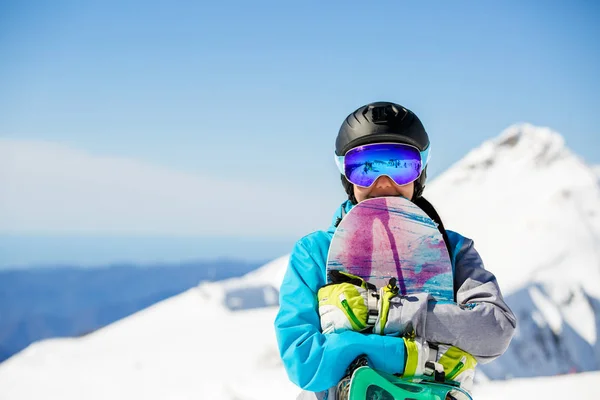 Foto einer jungen Touristin mit Helm, die mit dem Snowboard in der Hand vor dem Hintergrund von Schneebergen in die Kamera schaut — Stockfoto