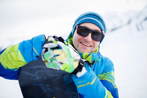 Retrato de hombre sonriente en gafas de sol con snowboard en manos sobre fondo de montañas nevadas —  Fotos de Stock