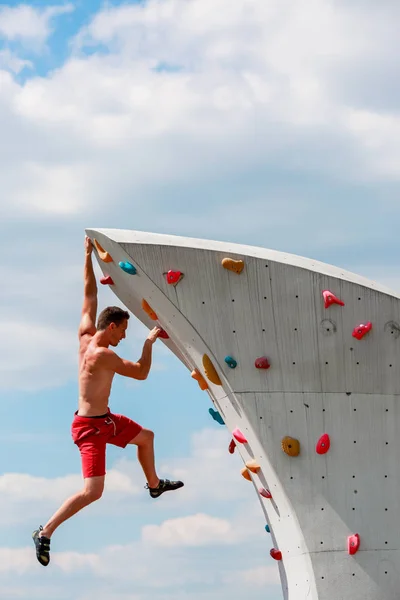 Photo of trained sports brunet in red shorts hanging on wall for rock climbing against blue sky with clouds
