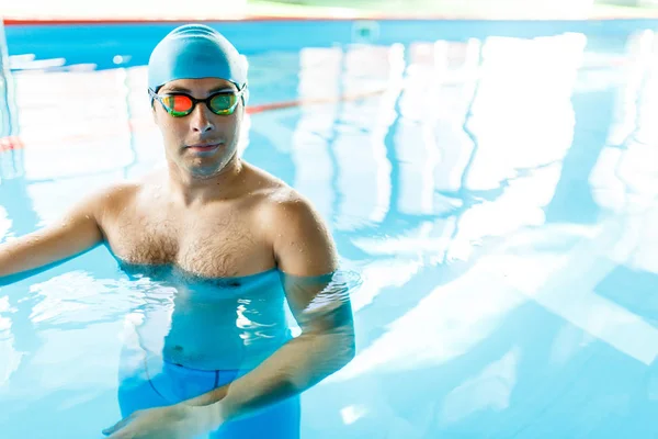 Image of athlete in blue rubber cap and glasses looking at camera in pool