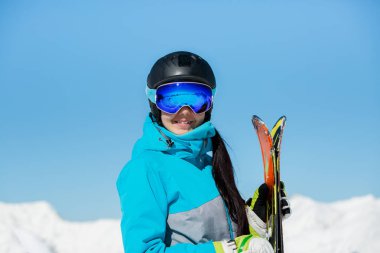 Photo of young smiling female athlete in helmet with skis in hand against blue sky and snowy hill clipart