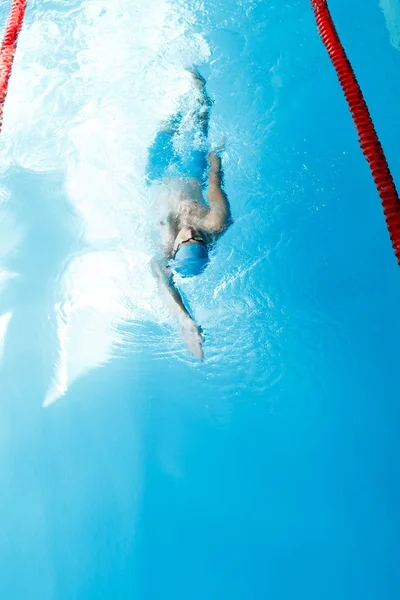 Foto op de top van sportieve jongeman in blauwe cap op rug in het zwembad zwemmen — Stockfoto