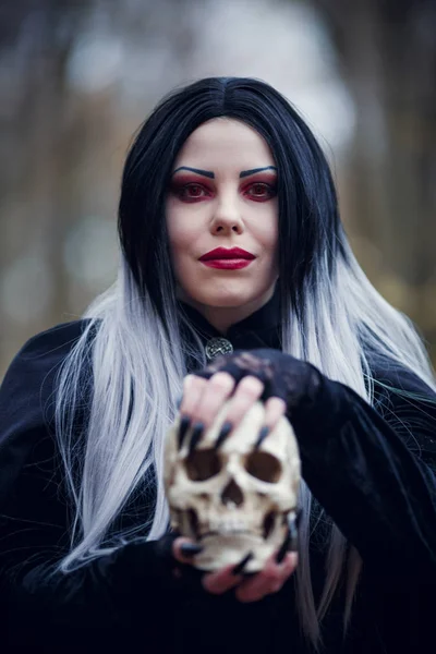 秋の森で手に頭蓋骨と黒マントの魔女女性のポートレート — ストック写真