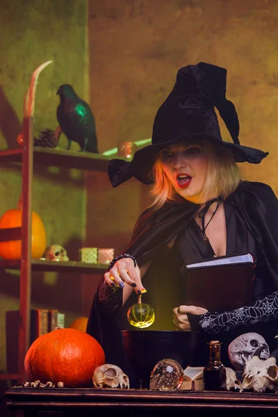 Foto de bruja en sombrero negro con libro de hechizos en la mesa con olla, calabaza —  Fotos de Stock