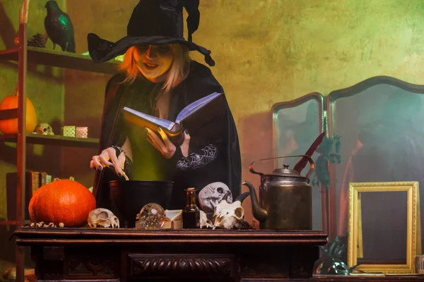 Foto de bruja en sombrero negro leyendo hechizo sobre olla con vapor verde — Foto de Stock