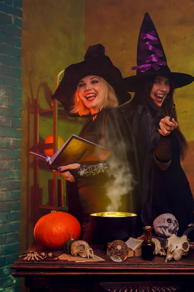 Foto de brujas sonrientes en sombreros negros con libro y caldero mágico en la mesa con calabaza y calaveras —  Fotos de Stock