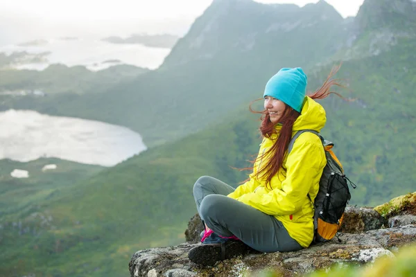 Изображение счастливой туристки с рюкзаком, сидящей на вершине горы на фоне живописного пейзажа — стоковое фото