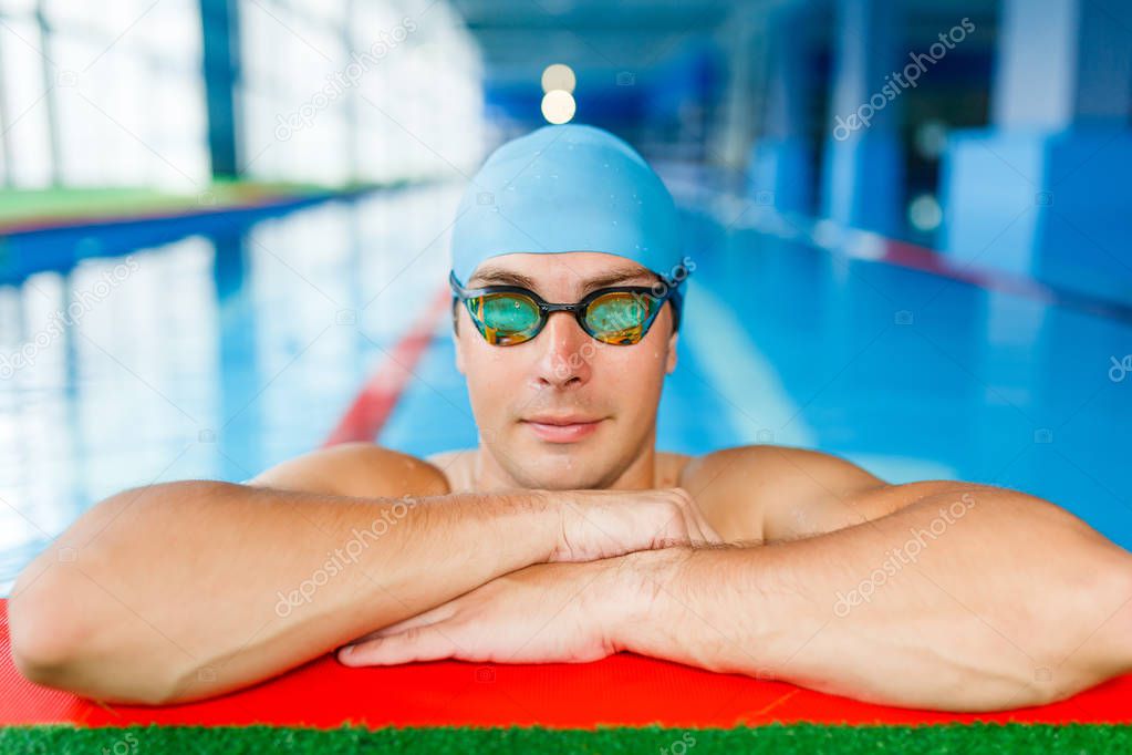 Portrait of athlete swimmer in blue cap, looking at camera at side in swimming pool