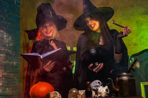 Portrait de sorcières souriantes en chapeaux noirs avec livre et chaudron magique à table avec citrouille et crânes — Photo