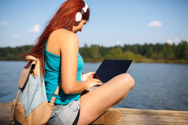 Imagen de mujer joven con auriculares en auriculares con mochila y portátil en manos sentadas en la orilla del río — Foto de Stock