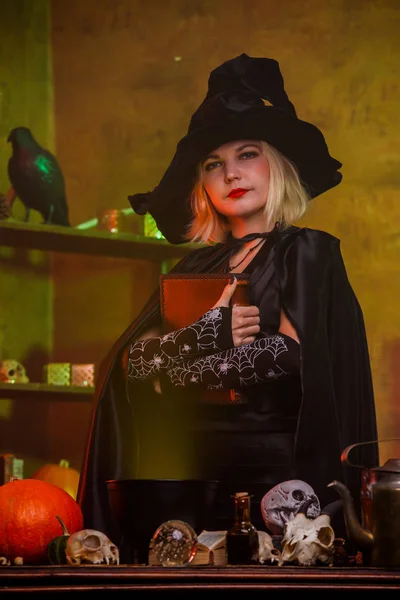 Photo de sorcière en chapeau noir avec livre de sorts à table avec pot, citrouille — Photo