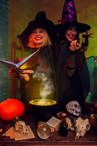Photo de sorcières souriantes dans des chapeaux noirs avec livre et chaudron magique à table avec citrouille et crânes — Photo