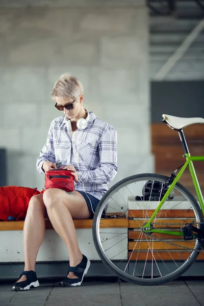 Foto einer jungen Blondine mit Kopfhörern, die auf einer Bank neben einem grünen Fahrrad sitzt — Stockfoto