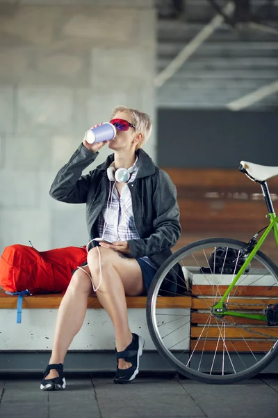 Фото молодой женщины, пьющей из стекла, сидящей на скамейке рядом с зеленым велосипедом — стоковое фото