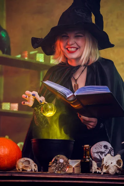 Foto van lachende blond heks in zwarte hoed met boek potgrond potion in pot — Stockfoto