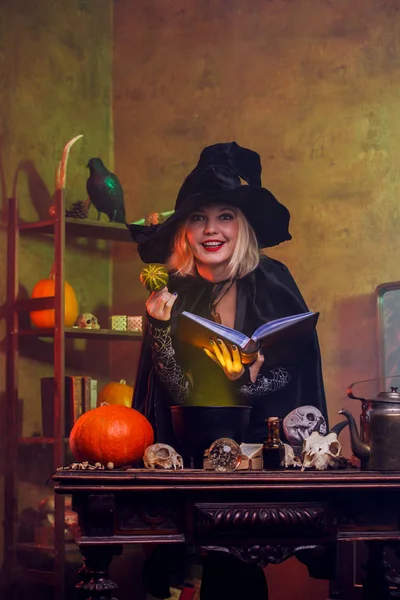 Image de sorcière blonde souriante en chapeau noir avec livre et citrouille dans les mains — Photo