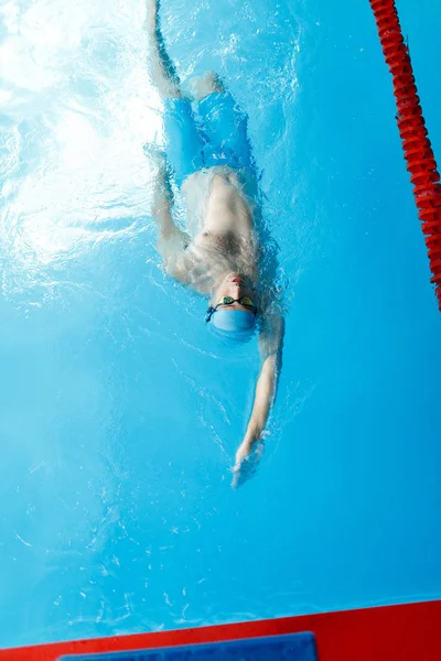 Foto op de top van jonge sporter in blauwe cap op rug in het zwembad zwemmen — Stockfoto