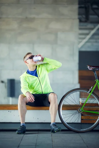 Foto de hombre en gafas de sol bebiendo de la glasse sentado en el banco cerca de bicicletas —  Fotos de Stock