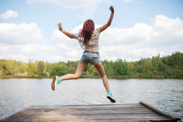 Photo du dos d'une jeune femme sautante sur un pont en bois au bord d'une rivière — Photo