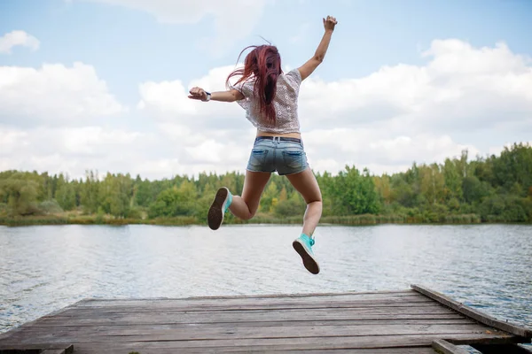 Image from back of young jumping woman on wooden bridge by river — Stock Photo, Image