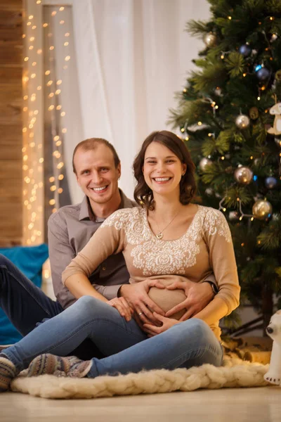 Bild des fröhlichen schwangeren Paares auf dem Hintergrund des geschmückten Neujahrsbaums — Stockfoto