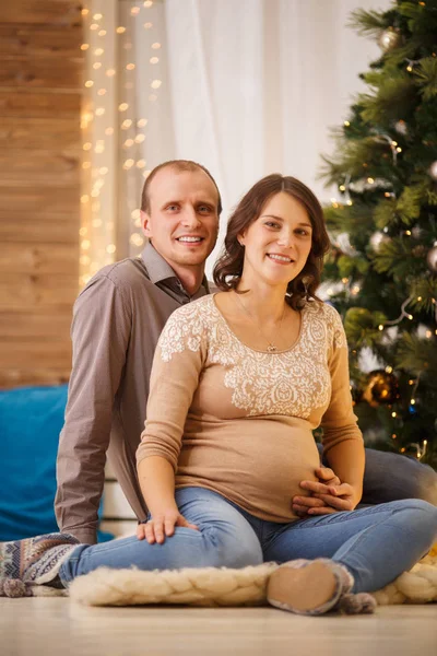 Зображення молодої вагітної пари на фоні прикрашеної новорічної ялинки — стокове фото
