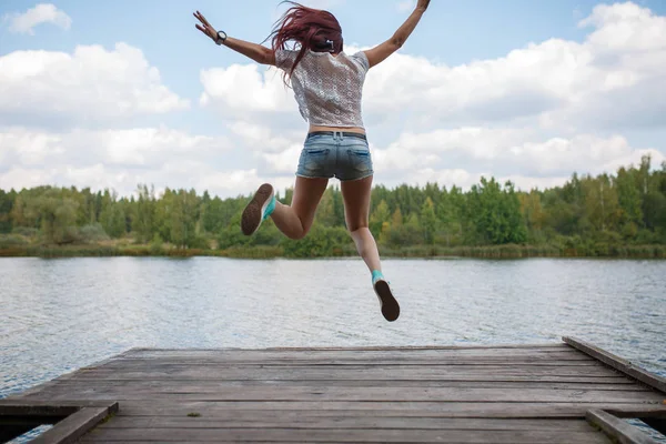 Photo from back of young jumping brunette on wooden bridge by river at day — Stock Photo, Image