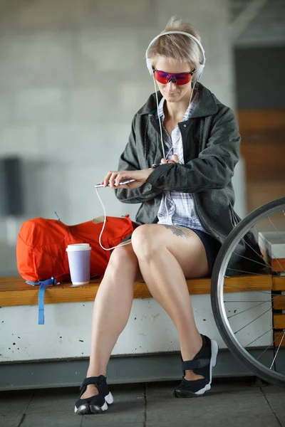 Φωτογραφία: νεαρή ξανθιά ν με το γυαλί που κάθεται στον πάγκο δίπλα στο καταπράσινο ποδηλάτων — Φωτογραφία Αρχείου