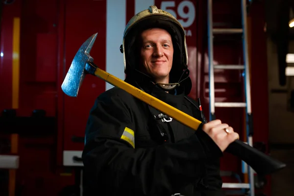 Foto de bombeiro em capacete de proteção com martelo olhando para a câmera contra o fundo do caminhão de bombeiros — Fotografia de Stock