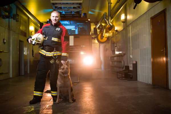 Foto de un bombero sosteniendo el casco en las manos y el perro en el fondo del camión de bomberos — Foto de Stock