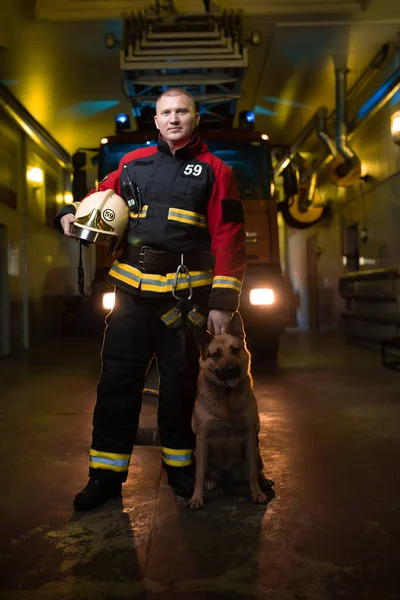 Bild eines Feuerwehrmannes mit Helm in der Hand und Hund auf dem Hintergrund eines Feuerwehrfahrzeugs — Stockfoto