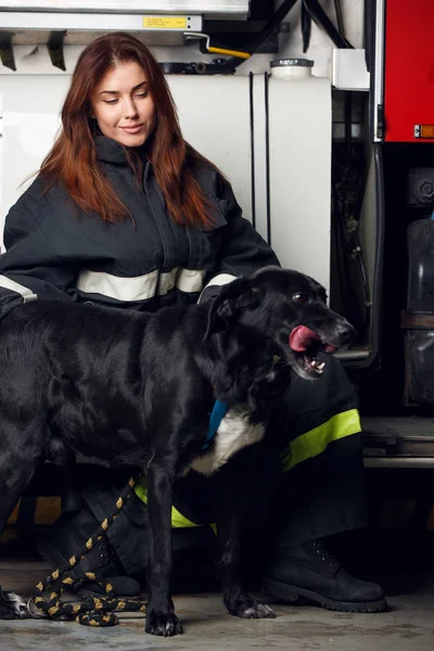Yangın kamyon arka plan üzerinde oturan siyah köpek ile kadın itfaiyeci portresi — Stok fotoğraf