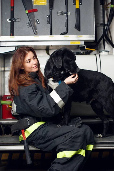 Photo de jeune femme pompier avec chien noir assis sur le fond du camion de pompiers — Photo