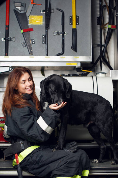 Image of young woman firefighter with black dog sitting on background of fire truck