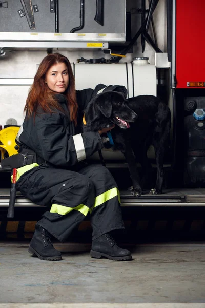 消防車の背景に座っている黒犬と若い女性消防士のイメージ — ストック写真