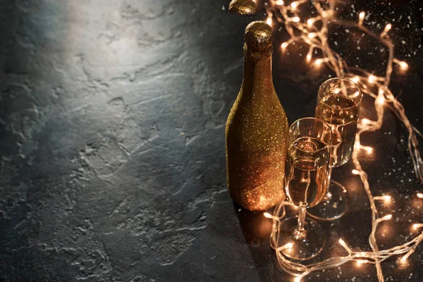 Photo of golden champagne bottle, two wine glasses, burning garland on black background — Stock Photo, Image