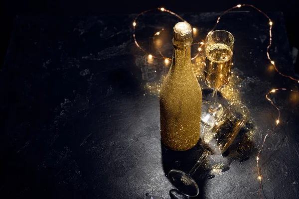 Photo romantique de bouteille de champagne doré, deux verres à vin, guirlande brûlante sur fond noir — Photo