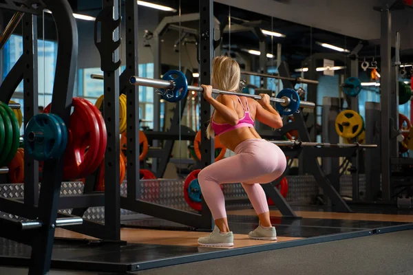 Foto de volta do atleta menina loira agachando-se com barbell em seus ombros no ginásio . — Fotografia de Stock