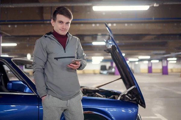 Uomo con il telefono in mano è in piedi vicino rotto auto blu con cappuccio aperto  . — Foto Stock